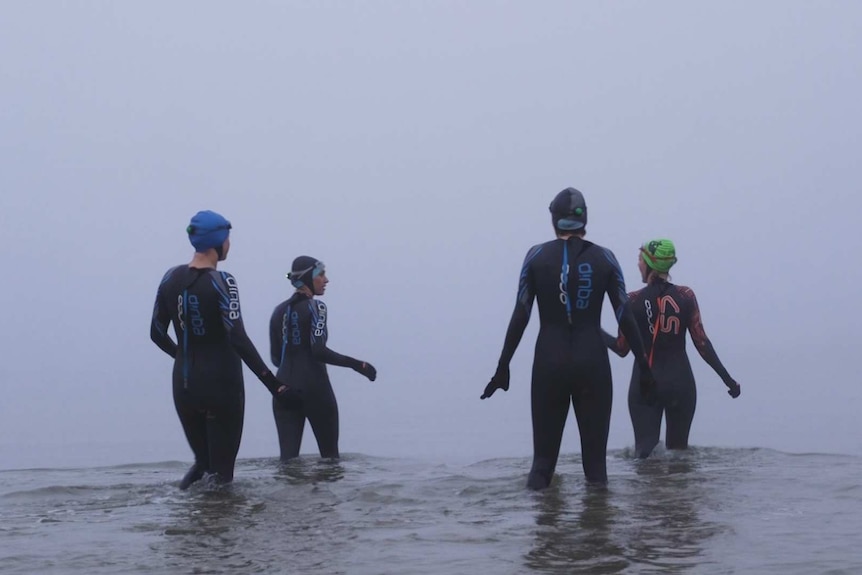 Four women in wetsuits stand in the ocean, blanketed by fog