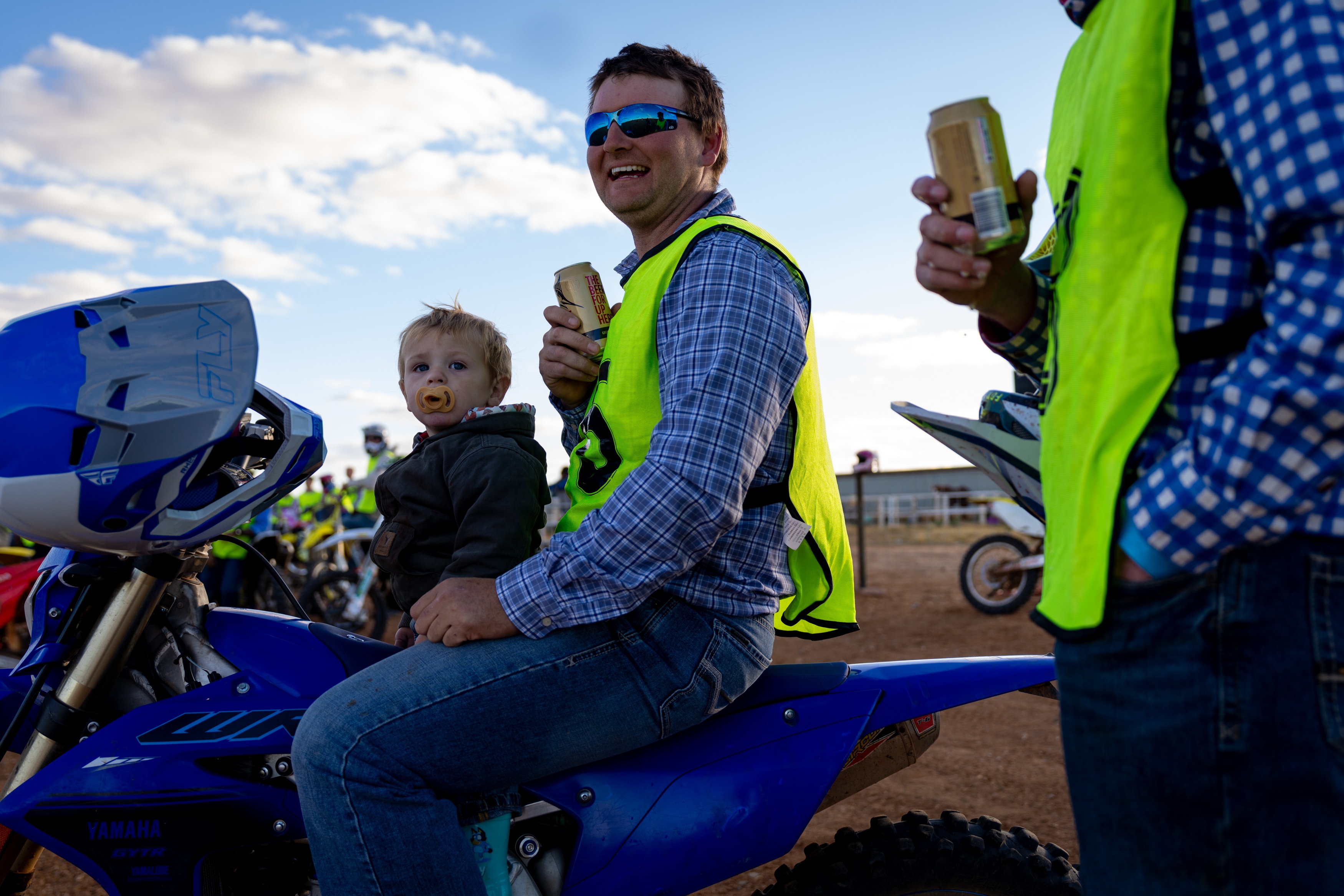 A man holding a beer sitting on a parked motorbike with his young son sitting infront of him