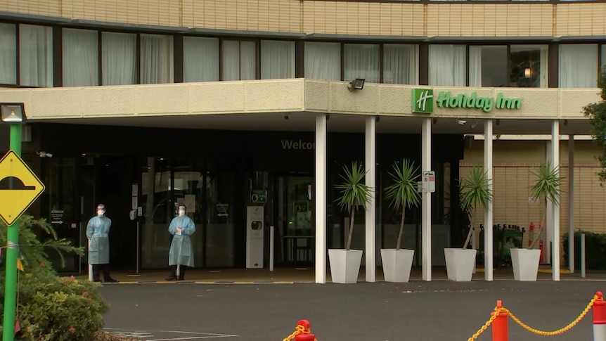 Two people in blue PPE gowns, masks and face shields stand outside the Holiday Inn hotel.