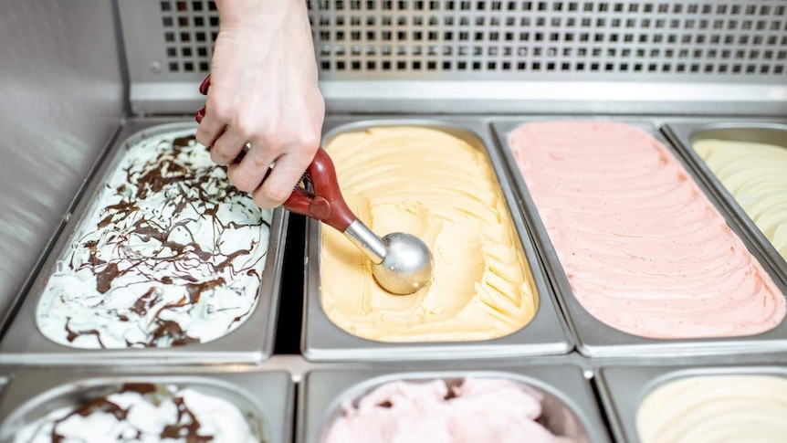 A scoop of ice cream being taken from a tub in a fridge of many flavours