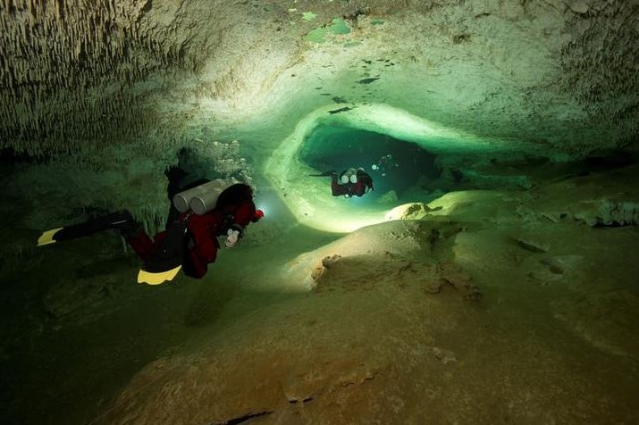 Two divers follow each other through an underwater cave lit up with a torch.