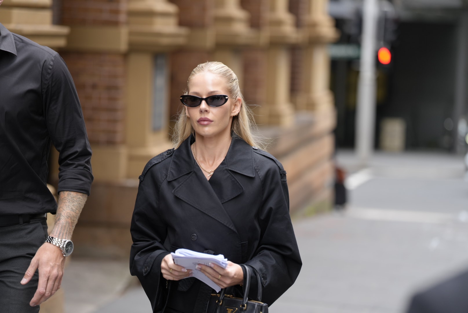 A young woman with blondee hair wearing black sunglasses and a black trench coat walks outside of court.