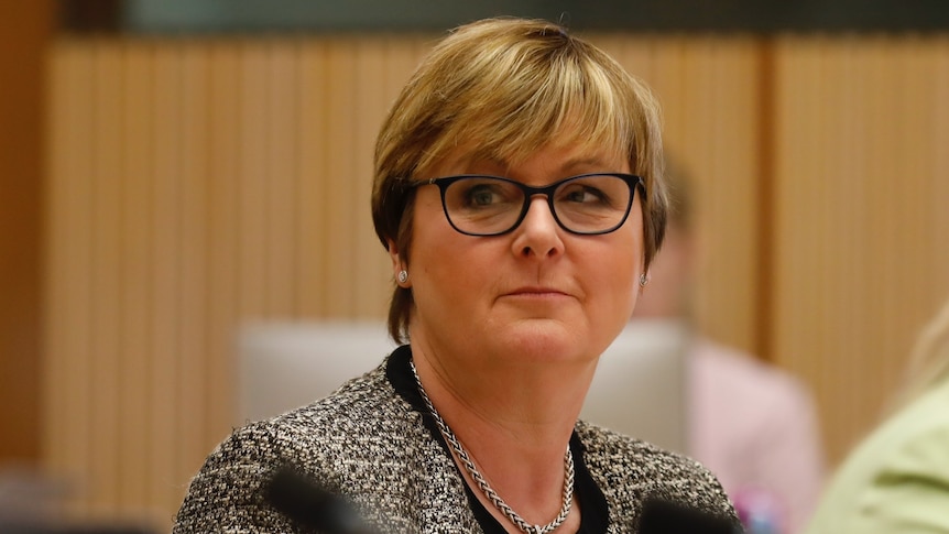 A woman with short blonde hair wearing glasses and a black and white blazer looking to the left