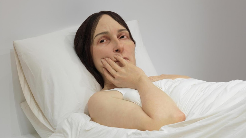 A hyperrealistic sculpture of a woman, with a hand raised to her face. She is lying in a bed, and appears concerned.