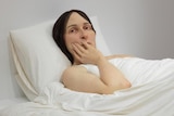 A hyperrealistic sculpture of a woman, with a hand raised to her face. She is lying in a bed, and appears concerned.