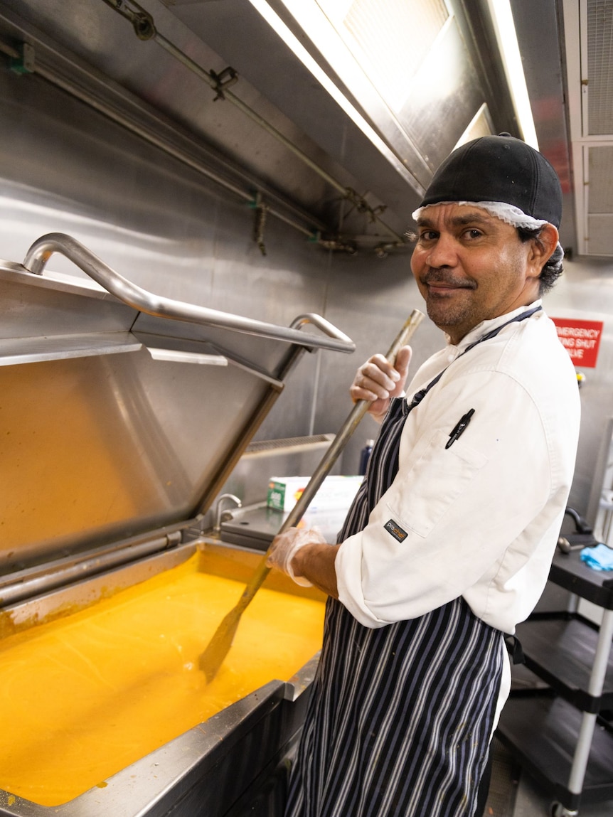 A man in a chef's uniform smiles at the camera and stirs a vat of yellow sauce.