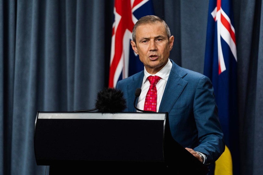Roger Cook wearing a blue suit, white shirt and red tie, standing at a lectern with microphones in front of flags.