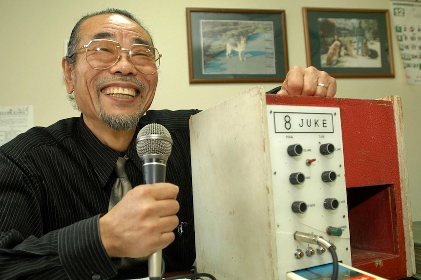  Daisuke Inoue, a Japanese man holding a microphone next to a box with a microphone coming out of it. 