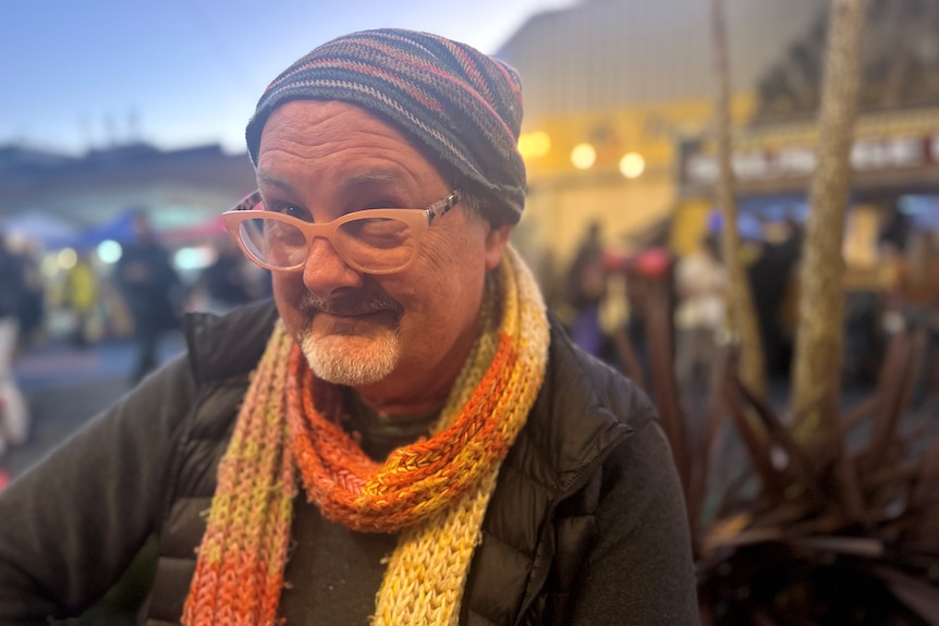 A man wears a colourful beanie and orange and yellow scarf. He looks over the top of his glasses