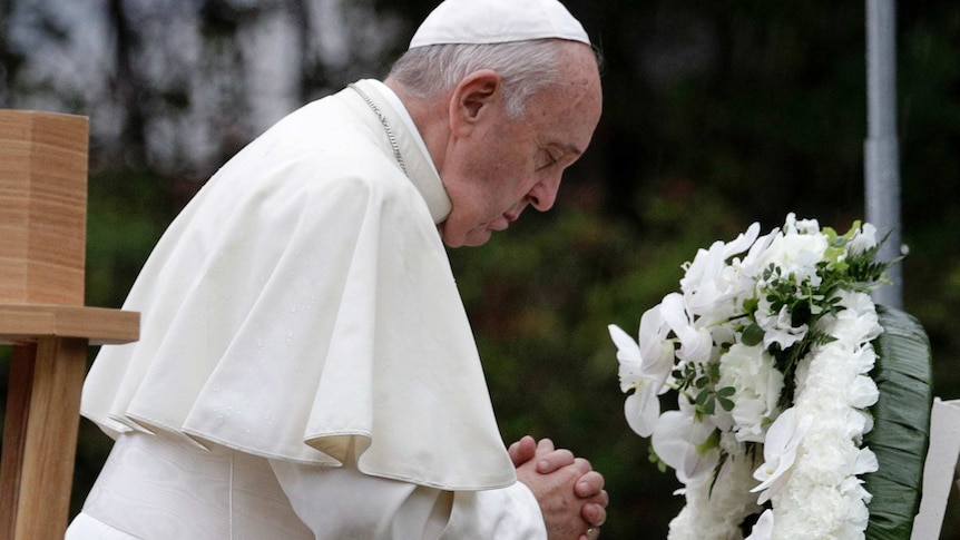 Pope Francis wearing papal robes bows his head, closes eyes, clasps hands in prayer in front of white wreath.
