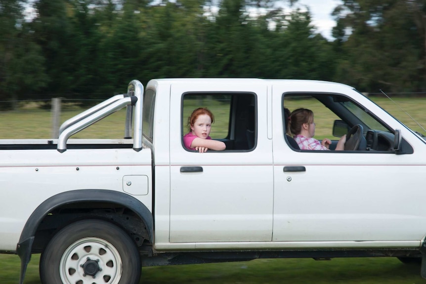 A girl drives a four-wheel drive on a farm with sister and friend in the car.