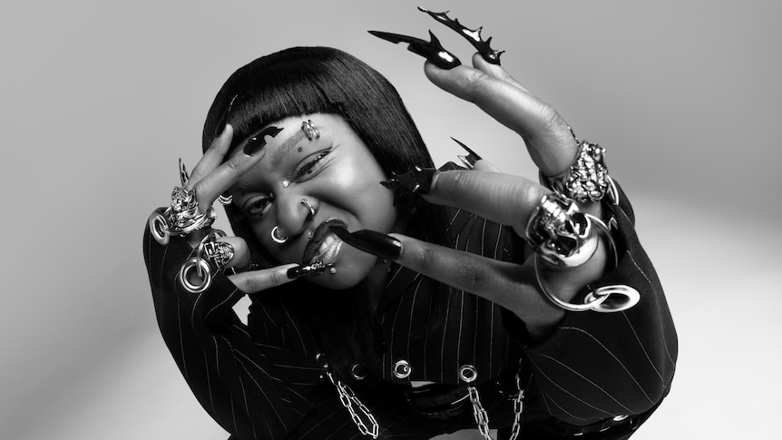 A woman with dark skin and hair holds painted fingernails towards the camera. She is wearing chunky jewellery and chains.
