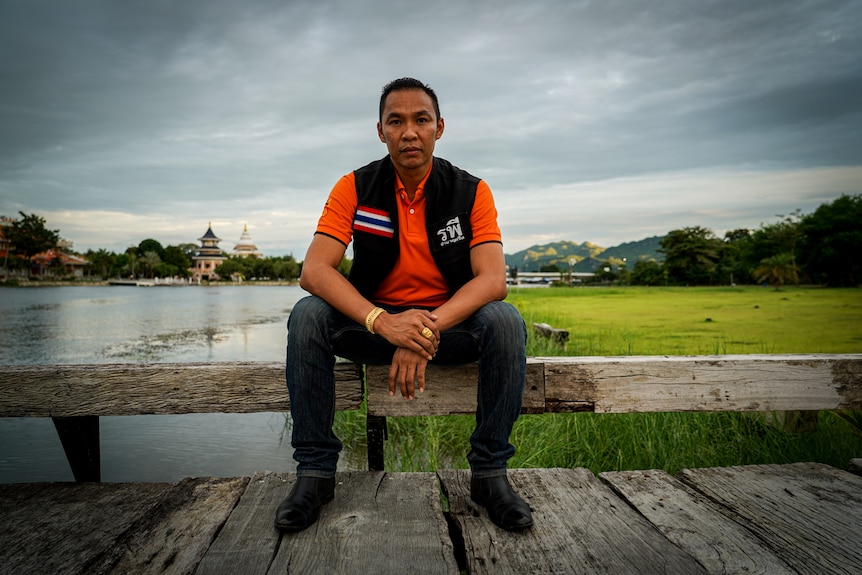 A man sitting down on a wooden ledge wearing an orange shirt and holding his arms.