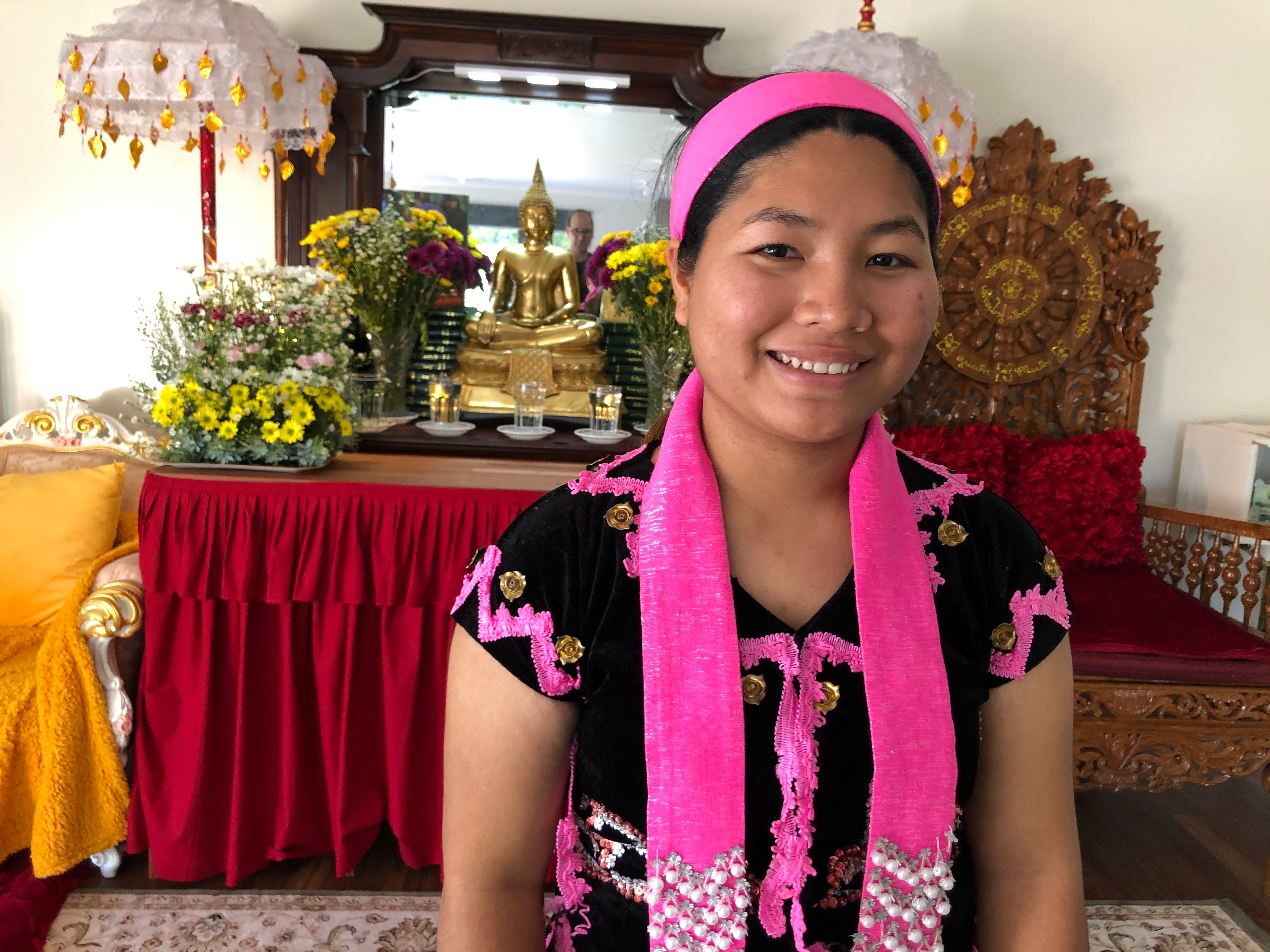 Ku Htee smiles and looks to the camera as she stands in front of a console. She wears pink and black traditional dress.