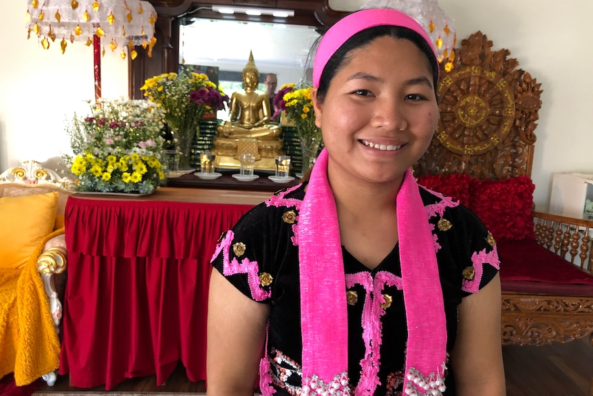 Ku Htee smiles and looks to the camera as she stands in front of a console. She wears pink and black traditional dress.