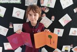 A young child in a school uniform holds up two coloured handmade cards