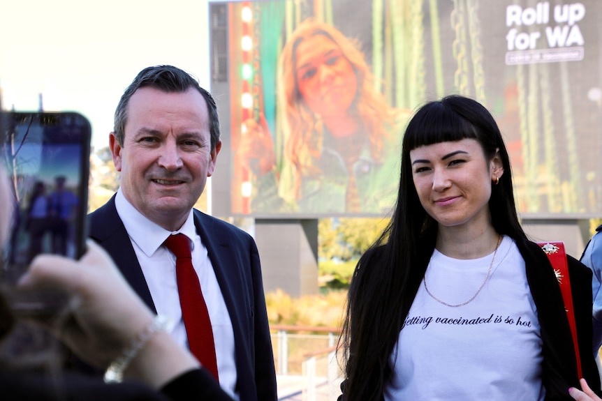 Someone takes a mobile photo of the Premier Mark McGowan and a woman with dark hair standing before a large video screen.