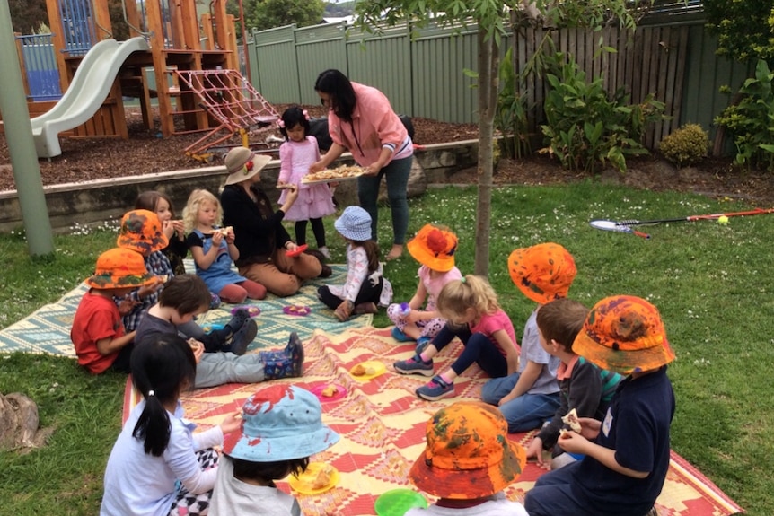 A group of children with orange hats sit on a rug outside 