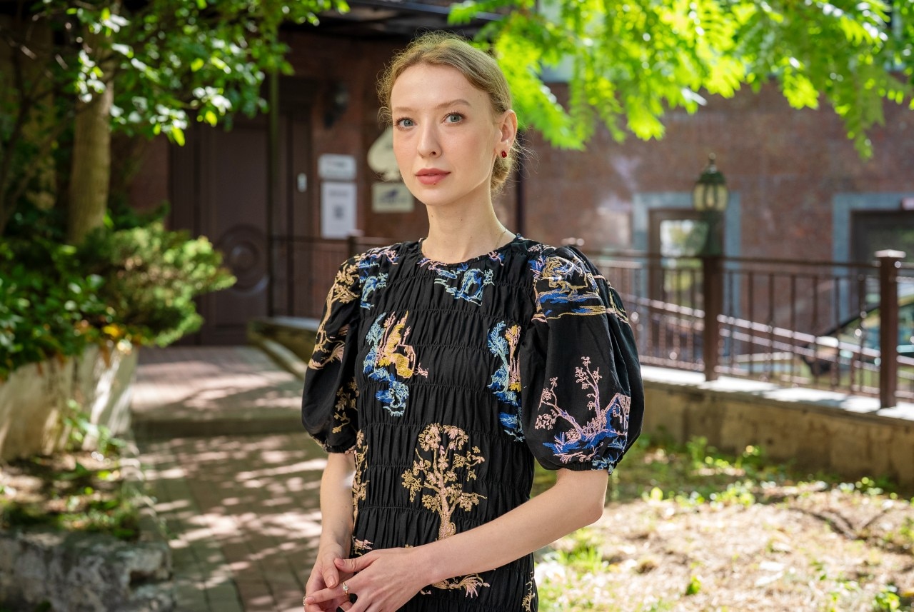 A woman wearing a patterned black dress standing outside looks at the camera with a serious expression.