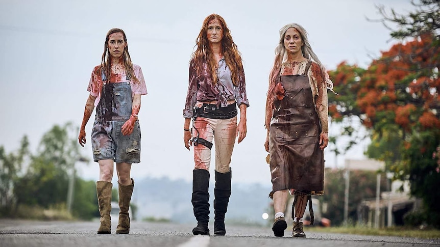 Three women walk on a bitumen road, covered in blood directly towards the camera