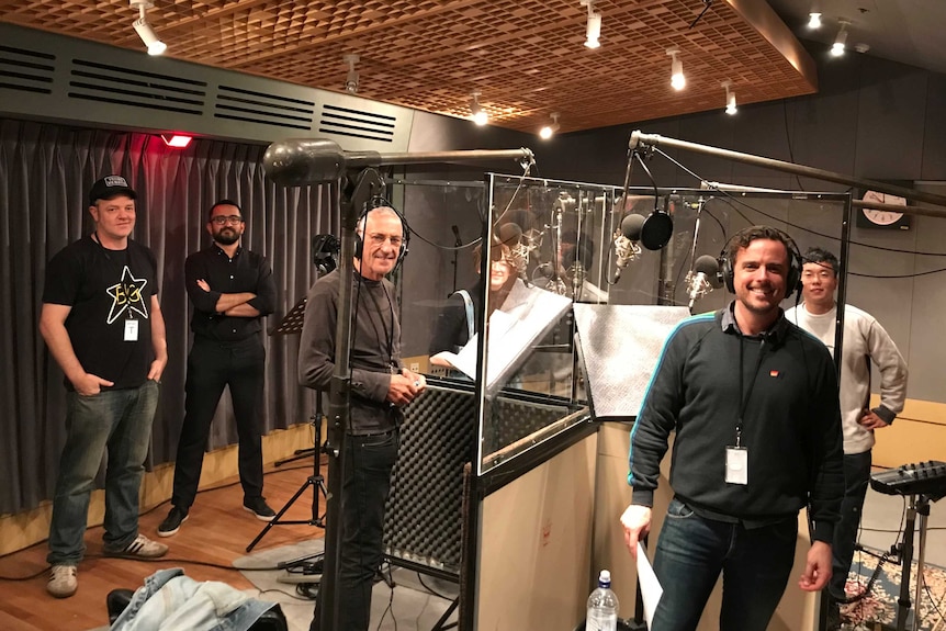 Cast standing at various microphones, separated by clear perspex screens in studio.