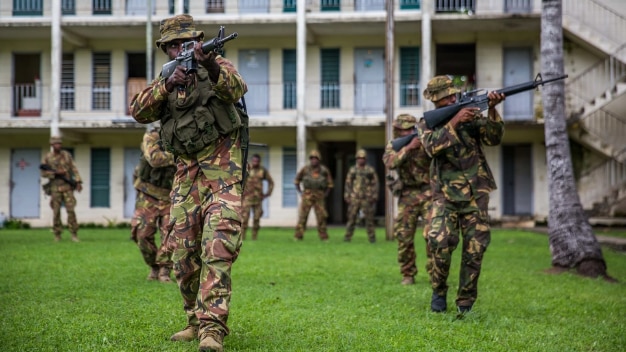 Officers in camouflage pointing guns moving toward camera during a training exercise with a building behind them