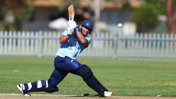 A male cricket player in a blue uniform strikes a cricket ball, on an oval