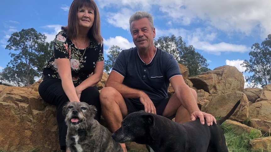 A smiling couple sit under a slightly cloudy blue sky with their dogs.
