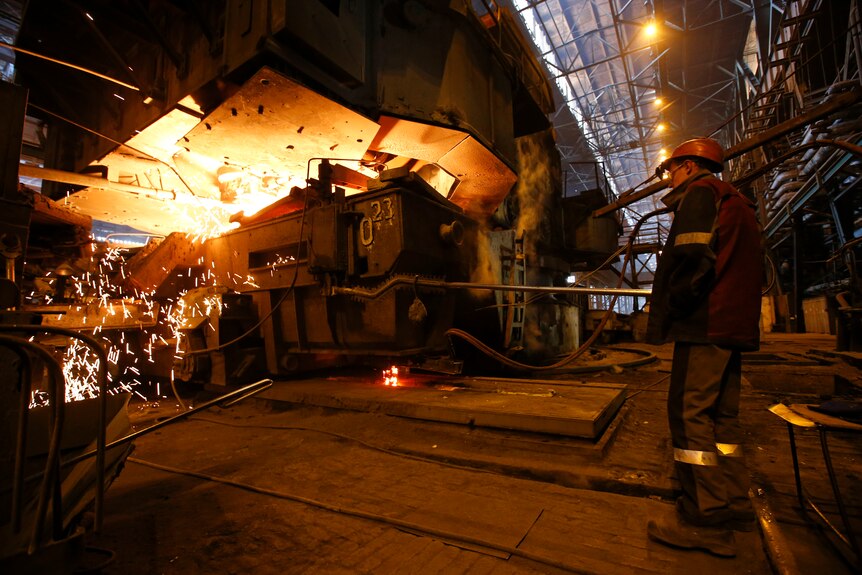 A worker stands next to a metallurgical machine in a factory. 