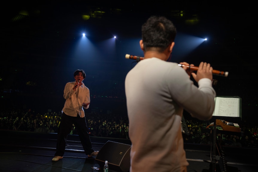 Un joven chino golpea un micrófono mientras otro toca la flauta en un escenario.