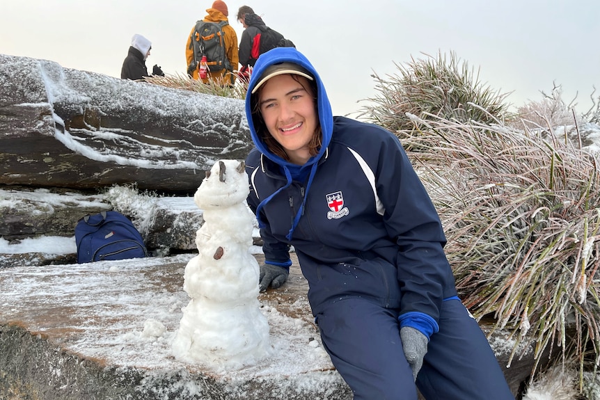 A smiling young man sits on a rock next to a snowman.