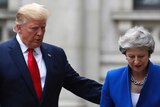 Trump walks in a suit wearing a red tie with his hand on Theresa May's back, she's wearing blue.