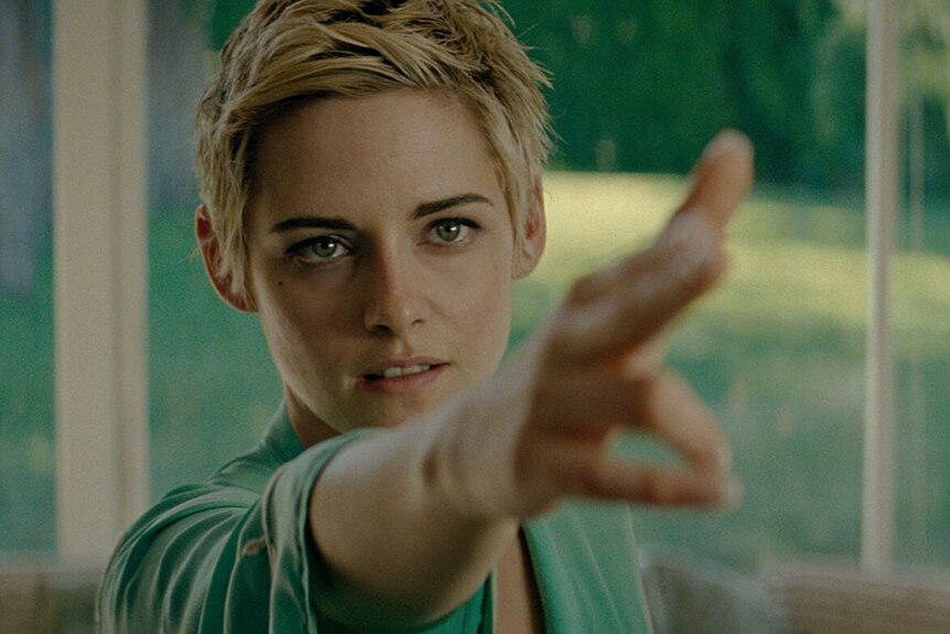 Kristen Stewart with short blonde pixie cut pointing hand towards right of frame with her fingers as gun.