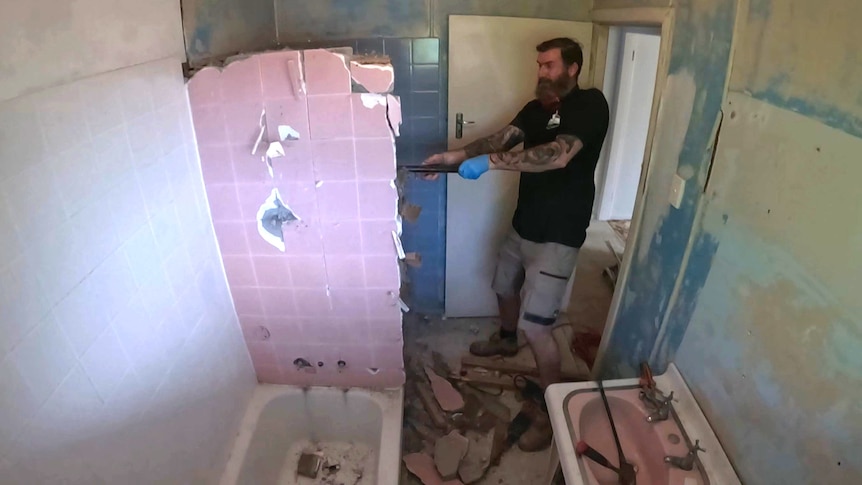 A wide angle shot of a man swinging a sledgehammer to get rid of a tiled wall in his bathroom 