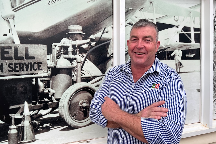 A man folds his arms and stands in front of a large historic photograph of an aeroplane.