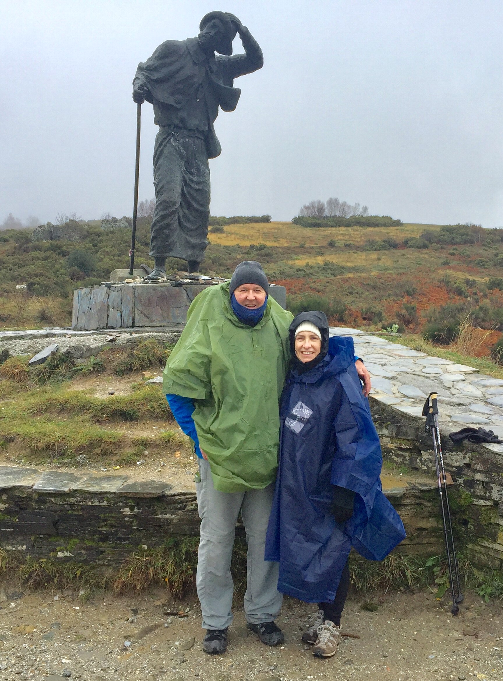 A man and woman in rain jackets stand next to a sculpture in the countryside.