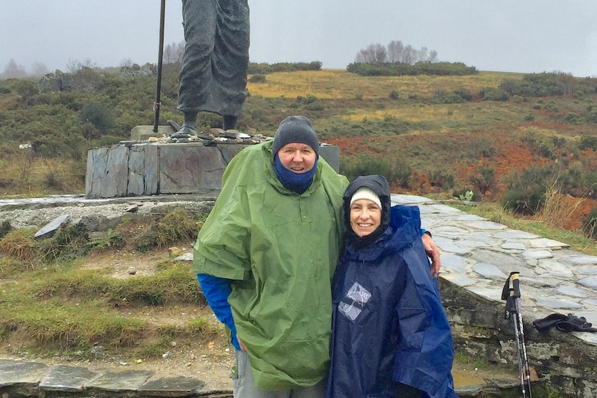 A man and woman in rain jackets stand next to a sculpture in the countryside.