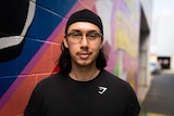 Young man leaning against a pink and purple multi coloured wall looking at the camera wearing glasses and half smiling