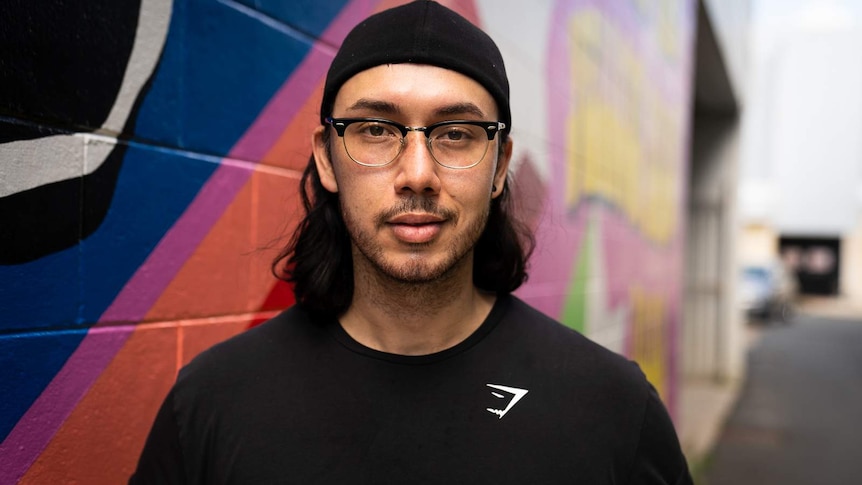 Young man leaning against a pink and purple multi coloured wall looking at the camera wearing glasses and half smiling