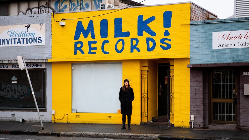 Courtney is wearing a coat and a beanie. She stands in front of a yellow shopfront that reads "MIlk! Records"
