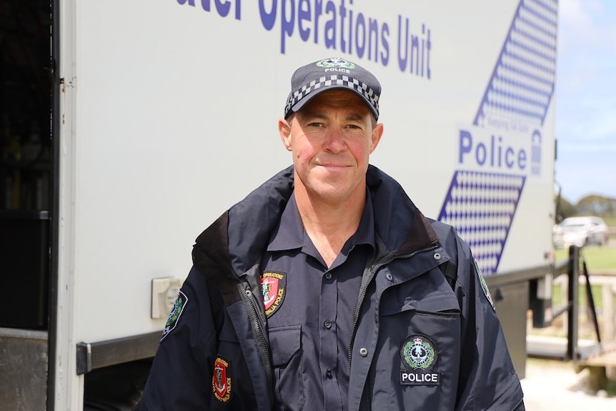 A man in a police uniform including cap stands in front of a police van that reads "operations unit".