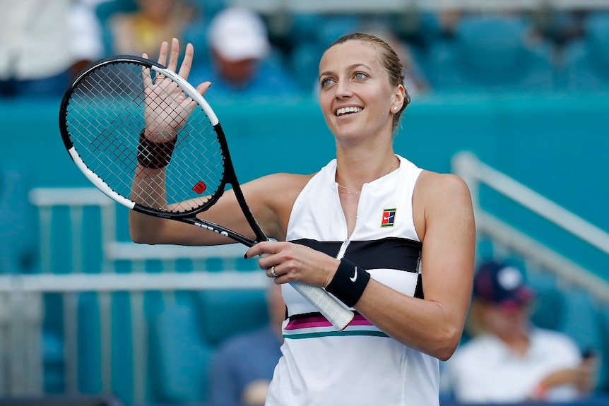 A female tennis player smiles as she looks up to the crowd from the court and claps her right hand against a tennis racquet.