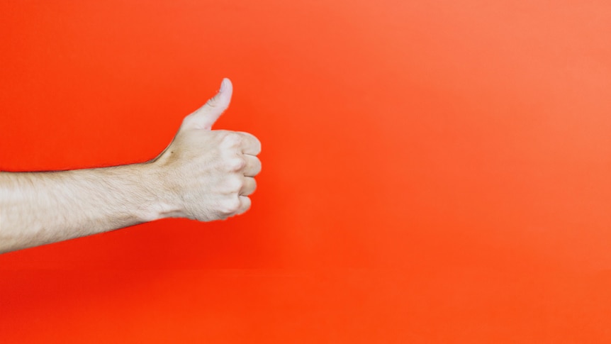 A hand gives the 'thumbs up' sign, orange wall in background.