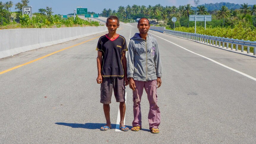 Two men stand on a giant empty freeway lined with palm trees