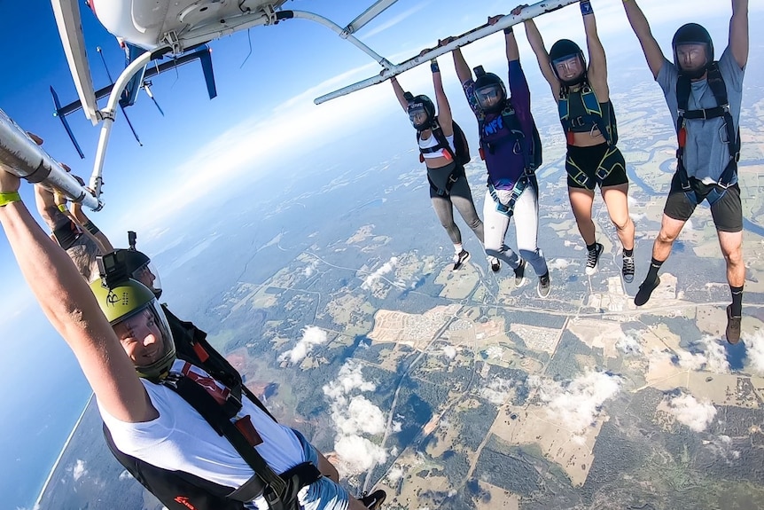 five skydivers hang by their arms under a helicopter hundreds of feet above the ground