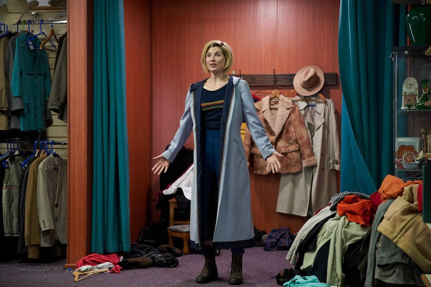 A woman in long coat, boots and high-waisted pants, shows off her outfit in a changing room filled with other clothes.