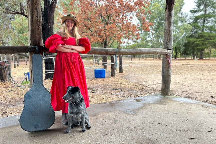 A woman wearing a red gown, standing on a farm with a blue heeler dog and a guitar case.