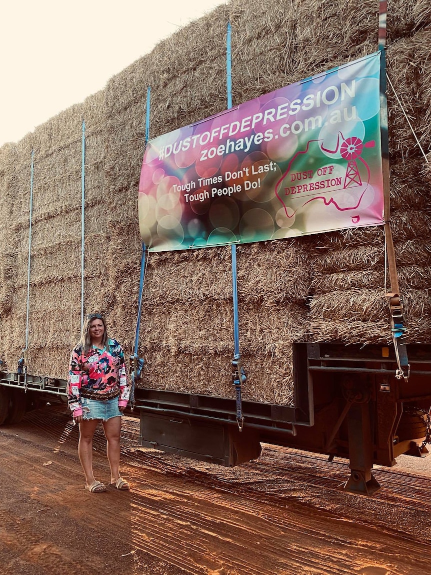 Zoe Hayes stands in front of truck loaded with hay and a sign with Dust off Depression logo.