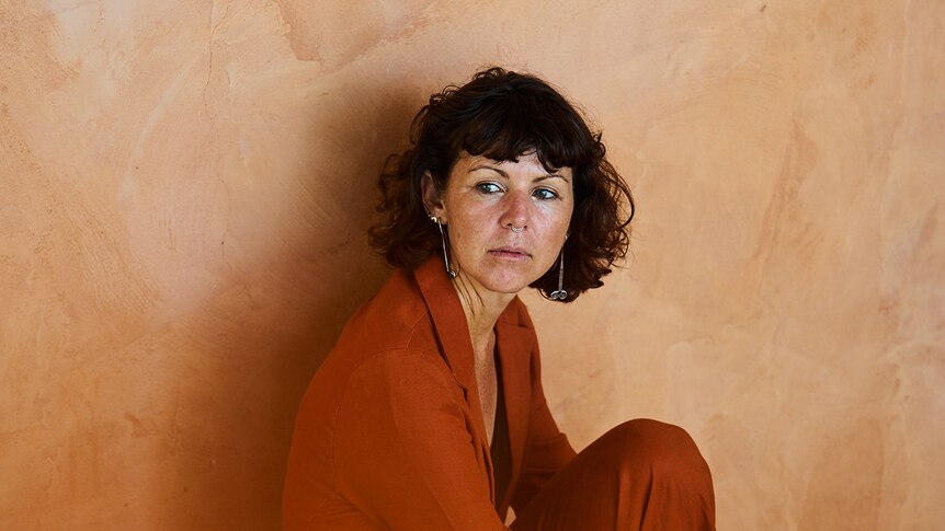 Tanya Ransom wears a burnt orange suit and sits in front of a terra cotta coloured wall