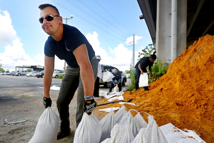 A man bends down to pick up sandbags as people in the background fill bags from a big pile of sand